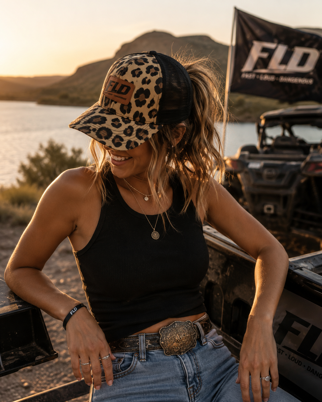 Woman wearing an FLD leopard ponytail trucker hat outdoors at sunset beside an off-road vehicle and lake.